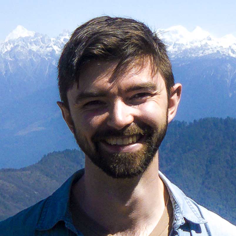 A portrait of Connor with dark brown hair and eyes, a short-kept beard, and with a backdrop of the Himalaya mountains